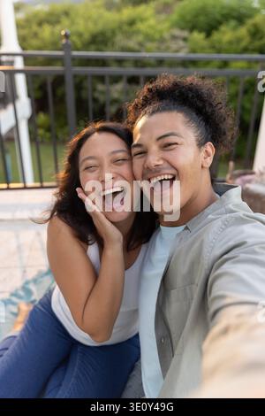 Diverse couple laughing and taking selfie on tiled balcony holding smartphone, woman in white tank Stock Photo