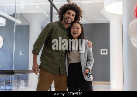 Diverse coworkers hugging near glass partition in office lobby holding scissors beside balloons. Collaboration, diversity, professionalism, celebration, modern, teamwork, positive || Model released Stock Photo