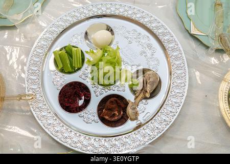 Traditional Passover seder plate with symbolic foods including egg, celery, lettuce, charoset, bitter herb, and roasted bone arranged on ornate silver tray, top view on festive holiday table setting. Stock Photo