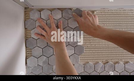 Worker's hands carefully installing grey marble-patterned hexagon mosaic tiles. Close-up on the process of laying backsplash flooring during a home im Stock Photo