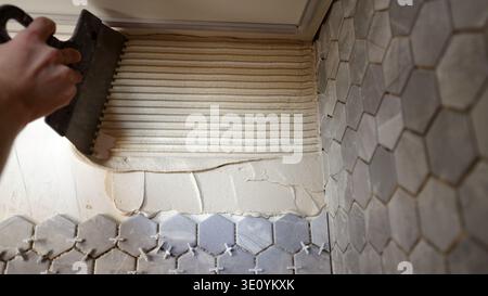 Close-up of a construction worker's hand using a notched trowel to spread mortar on a wall. Grey hexagonal tiles with plastic spacers are being instal Stock Photo