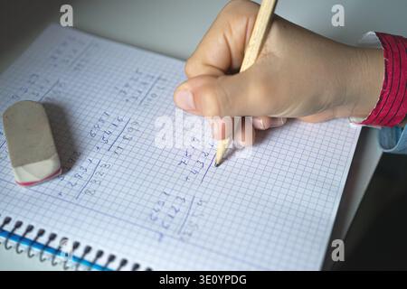 Close-up of a child's hand writing math problems in a graph paper notebook with a pencil. Concept of concentration, study, and school learning. Stock Photo