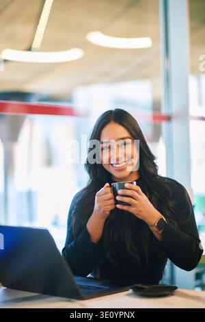 Smiling young Latina woman drinking coffee while working on laptop in modern cafe, happy female enjoying warm drink during remote work and relaxed wor Stock Photo