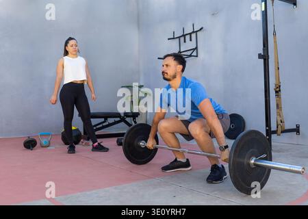 Focused male athlete performs a deadlift with a barbell while a female personal trainer observes his form in a modern outdoor gym setting. Promoting f Stock Photo