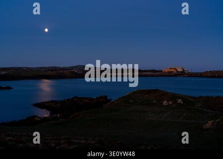 Bright moon reflecting on the calm water of Cemaes Bay with the industrial silhouette of Wylfa nuclear power station on the horizon Stock Photo