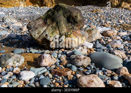 Close-up view of a large weathered boulder resting on a shoreline of multi colored pebbles and smooth stones at the base of coastal cliffs Stock Photo