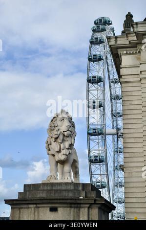 A carved stone lion stands sentinel on a plinth beside the London Eye ferris wheel and an ornate historic building, framed by blue sky and clouds to c Stock Photo