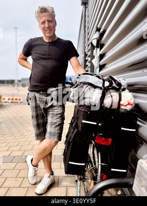 Middle aged man standing outdoors next to a loaded bicycle with panniers and bags, beside a camper van on a paved area. Casual travel and cycling scen Stock Photo