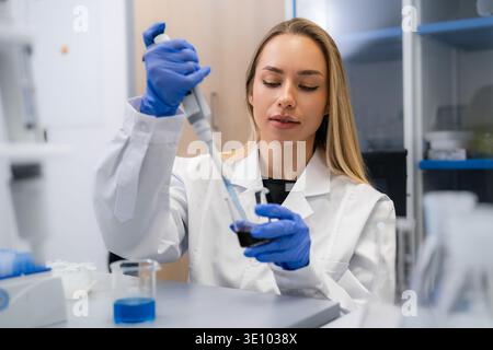 Technician using a micropipette to transfer liquid from one container to another at laboratory workstation. The workspace features various glassware and a clean, organized environment. Stock Photo