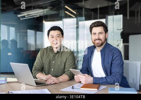 Two smiling male business partners, one asian and one caucasian, sitting at a desk with a laptop and digital tablet, symbolizing teamwork and global c Stock Photo