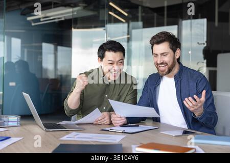 Two excited diverse businessmen celebrating a successful deal or project outcome, holding documents and expressing joy while working together at a mee Stock Photo