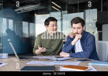 Mature asian business man offering support and empathy to a distressed male colleague in a modern office, showing teamwork and mental wellness at the Stock Photo