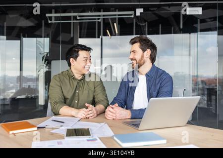 Two diverse male business partners discussing work and smiling while sitting at a table with papers and a laptop in a contemporary meeting room, showi Stock Photo