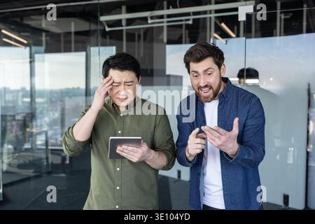 Two diverse male colleagues discussing business issues and expressing frustration over work challenges while looking at a digital tablet in a modern o Stock Photo