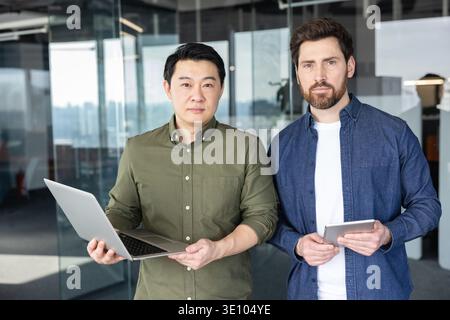 Diverse business partners standing confidently with a laptop and digital tablet, symbolizing teamwork, innovation, and modern technology in a contempo Stock Photo