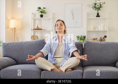 Senior elderly woman sitting on sofa in living room at home and meditating in lotus pose Stock Photo