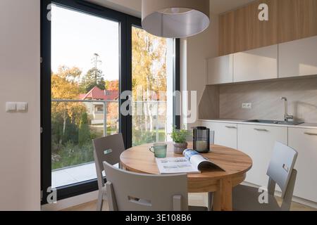 Modern apartment interior featuring a round wooden dining table with magazine and coffee. Kitchenette with white cabinets in the background. Large bal Stock Photo