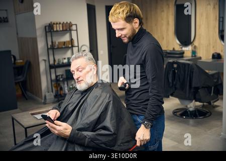 Mature man using smartphone while sitting in barbershop, talking with barber Stock Photo