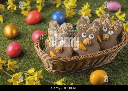 Bunny shaped Easter cookies with chocolate and carrot in a basket surrounded by colorful eggs on grass Stock Photo