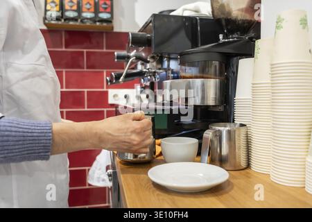 Barista wearing a white apron steaming milk in a metal jug, working at a professional espresso machine on a wooden counter with stacks of disposable c Stock Photo