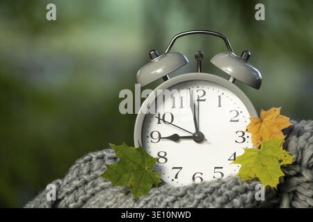 A close up of a classic alarm clock surrounded by colorful autumn leaves, resting on a soft, knitted scarf, with a blurred green background Stock Photo