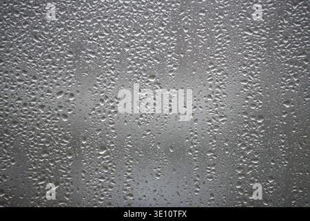Close-up view of water condensation droplets forming patterns on a foggy glass window, creating a textured and abstract background illuminated by ligh Stock Photo