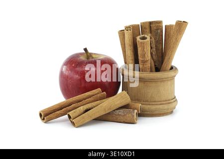 A vibrant red apple next to cinnamon sticks arranged in a wooden container, captured on a white background, conveying freshness and warmth Stock Photo