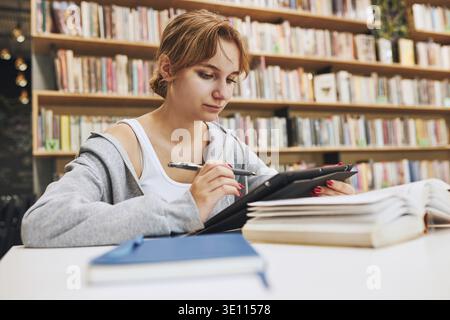Student learning in university library. Young woman writing essay and making notes using computer tablet. Focused student studying for college exams. Stock Photo
