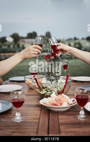 Couple making toast during summer outdoor dinner in a home garden. Hands holding wine glasses with red wine over table with dishes Stock Photo