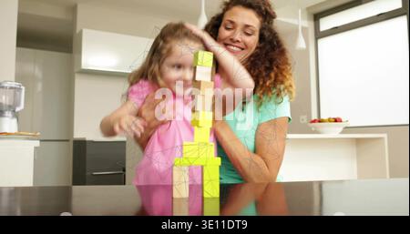 Guiding mom in mint, daughter in pink stacking blocks on glossy counter in kitchen reflecting tower Stock Photo