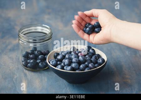 Woman putting freshly gathered blueberries from a jar into a small bowl. Table painted in blue in the background Stock Photo