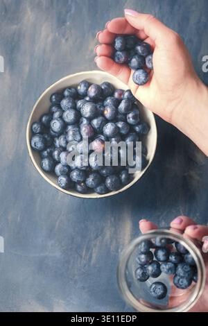 Woman putting freshly gathered blueberries from a jar into a small bowl. Table painted in blue in the background Stock Photo