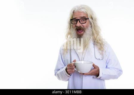 Studio shot of happy senior bearded man doctor smiling while holding coffee cup Stock Photo
