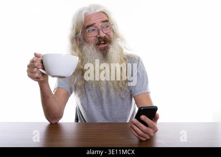Thoughtful happy senior bearded man smiling while holding mobile phone and raising coffee cup on wooden table Stock Photo