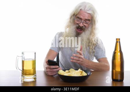 Studio shot of happy senior bearded man smiling while using mobile phone with beer and bowl of potato chips on wooden table Stock Photo
