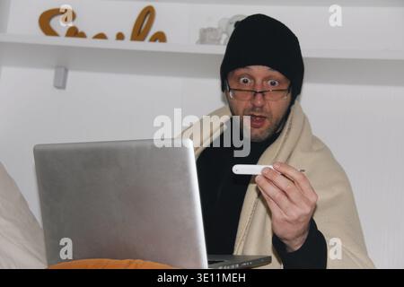 Man With Fever Wrapped in Blanket Checking Thermometer While Using Laptop at Home Stock Photo