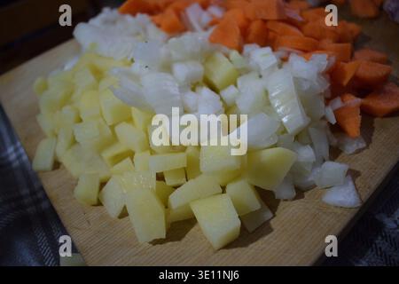 A top-down view of freshly diced carrots, onions, and potatoes neatly arranged on a wooden cutting board. Stock Photo