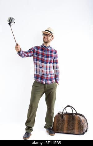 Full body shot of happy young man smiling while holding selfie stick and taking selfie picture with mobile phone ready for vacation Stock Photo