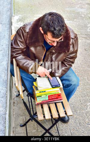 Young man sitting outside cafe with a pile of textbooks checking information notes on a smartphone - Saint-Gilles, Brussels, Belgium. Stock Photo