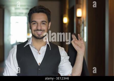 Young happy Indian man smiling while holding jacket in the corridors of hotel Stock Photo
