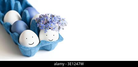Easter eggs with painted faces and flowers in a blue cardboard box on a white background. Copy space. Banner. Stock Photo