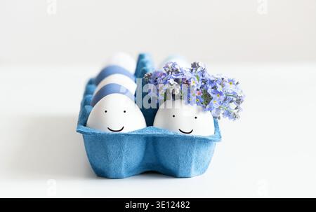 Easter composition with eggs in a blue cardboard box on a white background. Couple of funny eggs with painted faces and flowers. Stock Photo