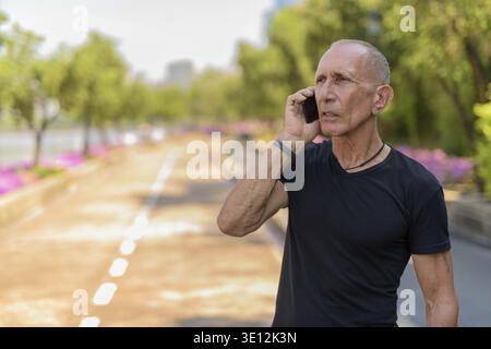 Thoughtful bald senior tourist man talking on mobile phone on the side of the street at peaceful park in Bangkok Thailand Stock Photo