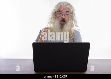 Studio shot of senior bearded man looking shocked while using laptop on wooden table Stock Photo