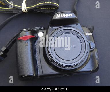 Nikon D3000 digital camera body with attached lens cap and black camera strap resting on a dark surface Stock Photo