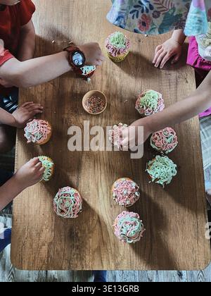 Group of children baking cupcakes, preparing ingredients, topping, sprinkles for decorating cookies. Kids cooking, working together in kitchen at home Stock Photo