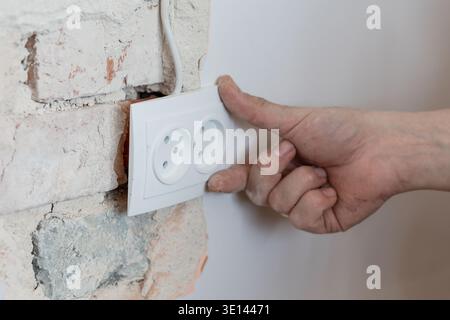 Electrician Installing White Double Socket Outlet On Exposed Brick Wall With Dusty Hand And Visible Wiring In Home Renovation Scene Stock Photo