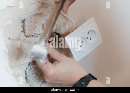 Electrician Hammering Wall Near White Dual Socket Outlet During Renovation Project Showing Exposed Brick Texture And Construction Tools In Action Stock Photo