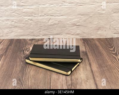Two professional black journals with elastic closures stacked on a rustic wooden surface against a white brick wall. Stock Photo