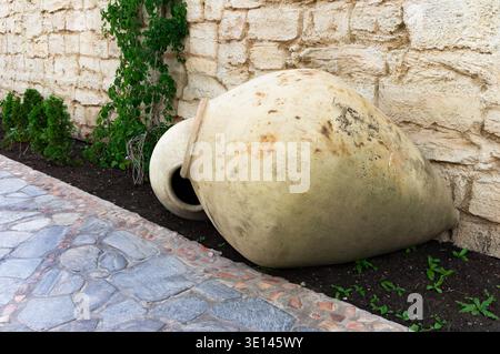 A large, brown, ceramic vase is sitting on the ground next to some plants. The vase is in the middle of a garden and is surrounded by green plants Stock Photo